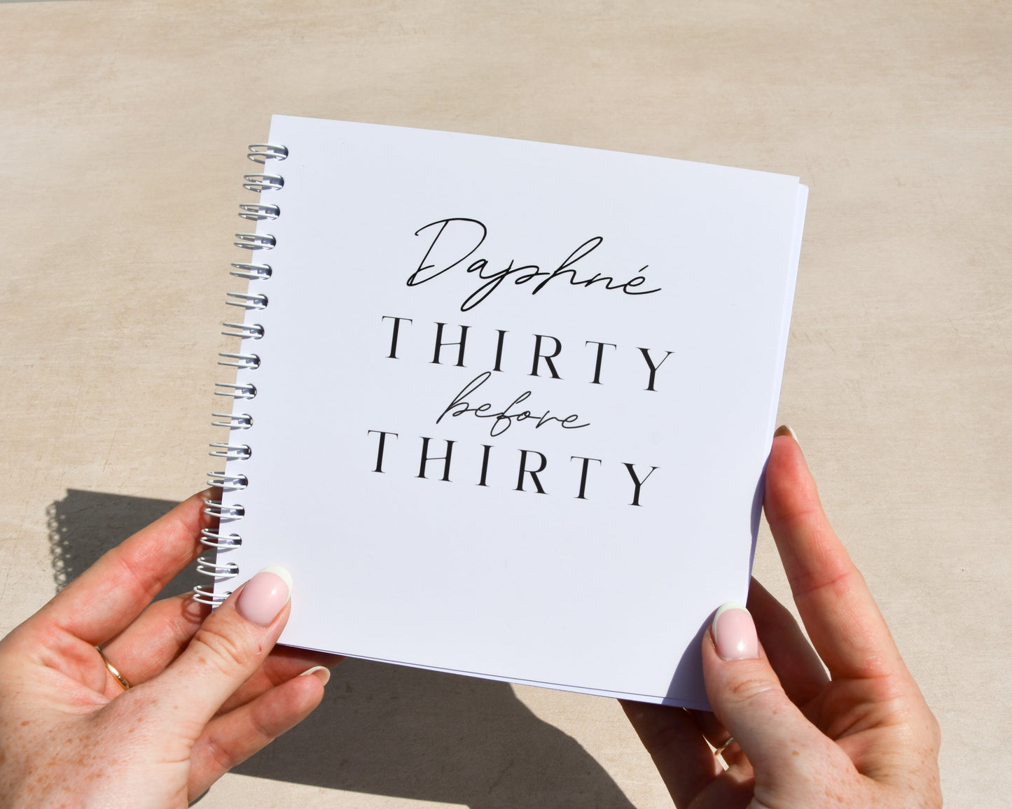 Person holding a notebook titled 'Daphne Thirty before Thirty' on a light surface.