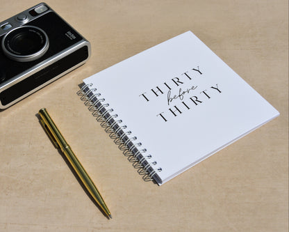 Black camera, gold pen, and notebook titled 'Thirty before Thirty' on a wooden surface