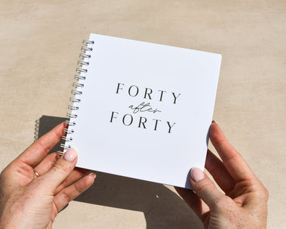 Person holding a spiral-bound notebook titled 'Forty After Forty' on a desk.