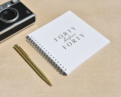 Camera, notebook with 'Forty before Forty' text, and pen on a beige surface
