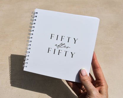 Hand holding a notebook titled 'Fifty after Fifty' on a light surface.