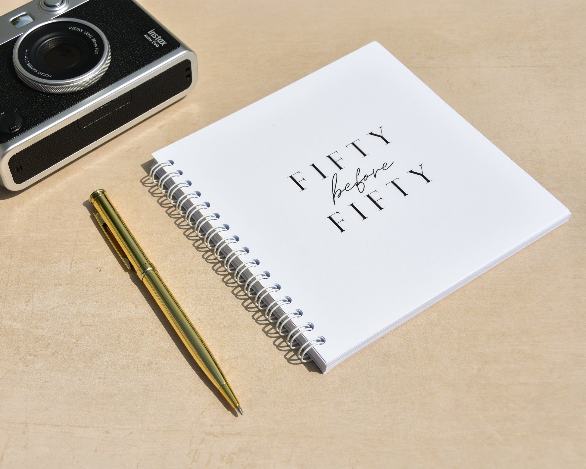 Camera, pen, and fifty before fifty notebook on a beige surface