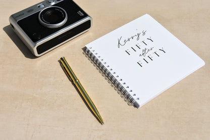 Camera, pen, and notebook with 'Kerry's Fiftieth Birthday' text on a beige surface