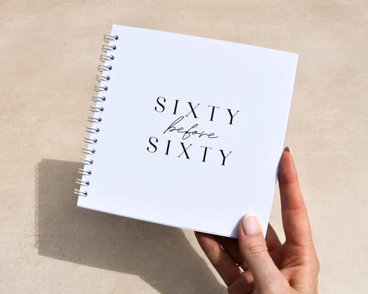 Hand holding a white notebook titled 'Sixty before Sixty' on a beige surface.