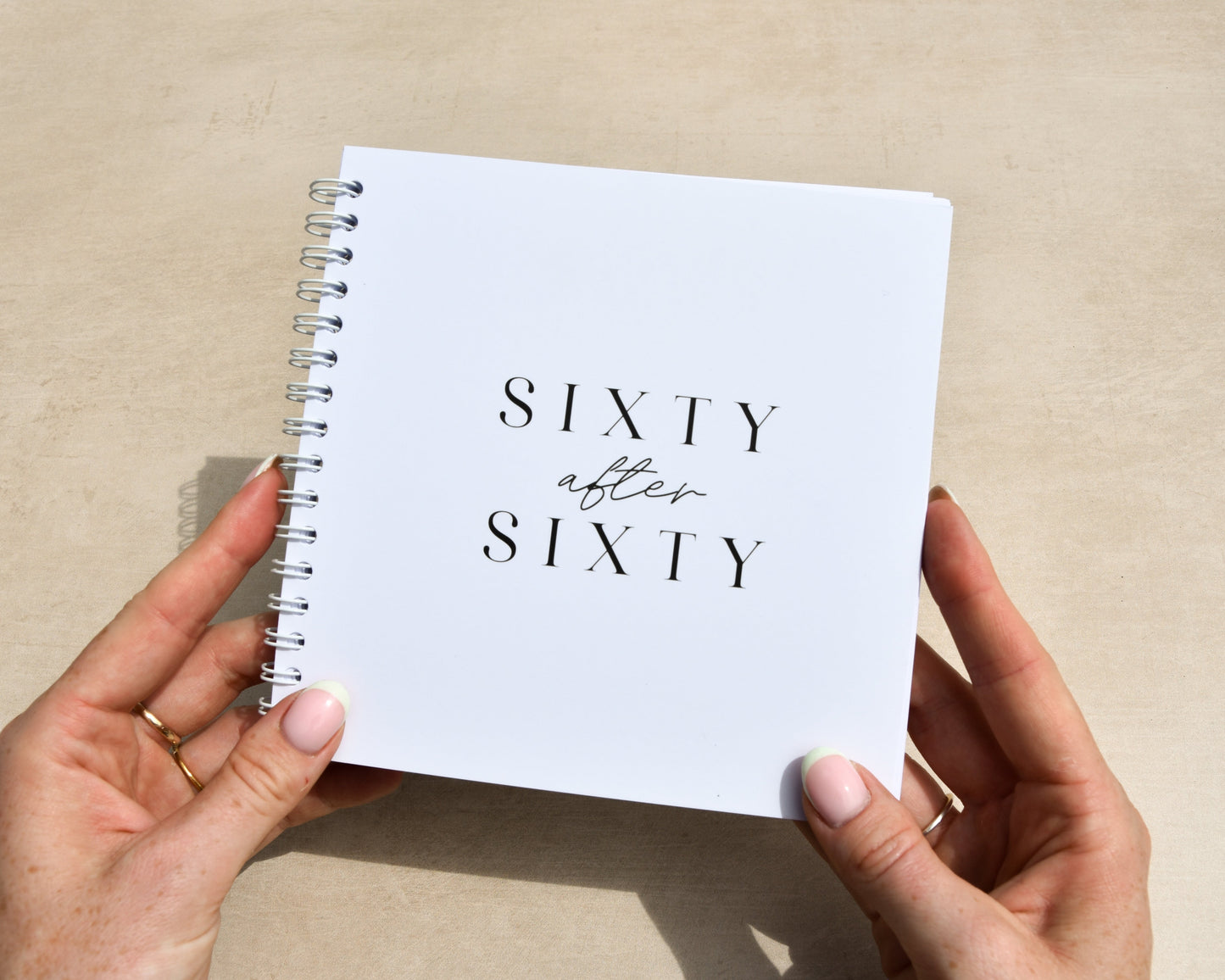 Person holding a small white notebook titled 'Sixty after Sixty' on a beige background