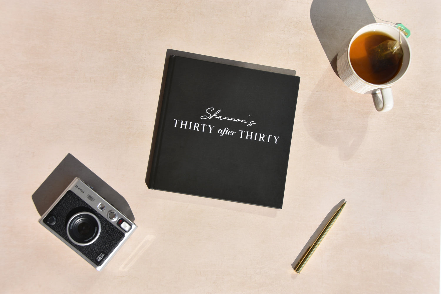 Luxury black hardback 30 After 30 journa with 'Shannon's Thirty after Thirty' text, vintage camera, pen, and mug on a beige surface.