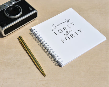 Spiral-bound notebook titled 'Laura's Forty after Forty' with a camera and pen on a beige surface