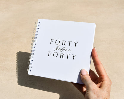 Hand holding a spiral-bound notebook with 'Forty before Forty' text on a beige surface.