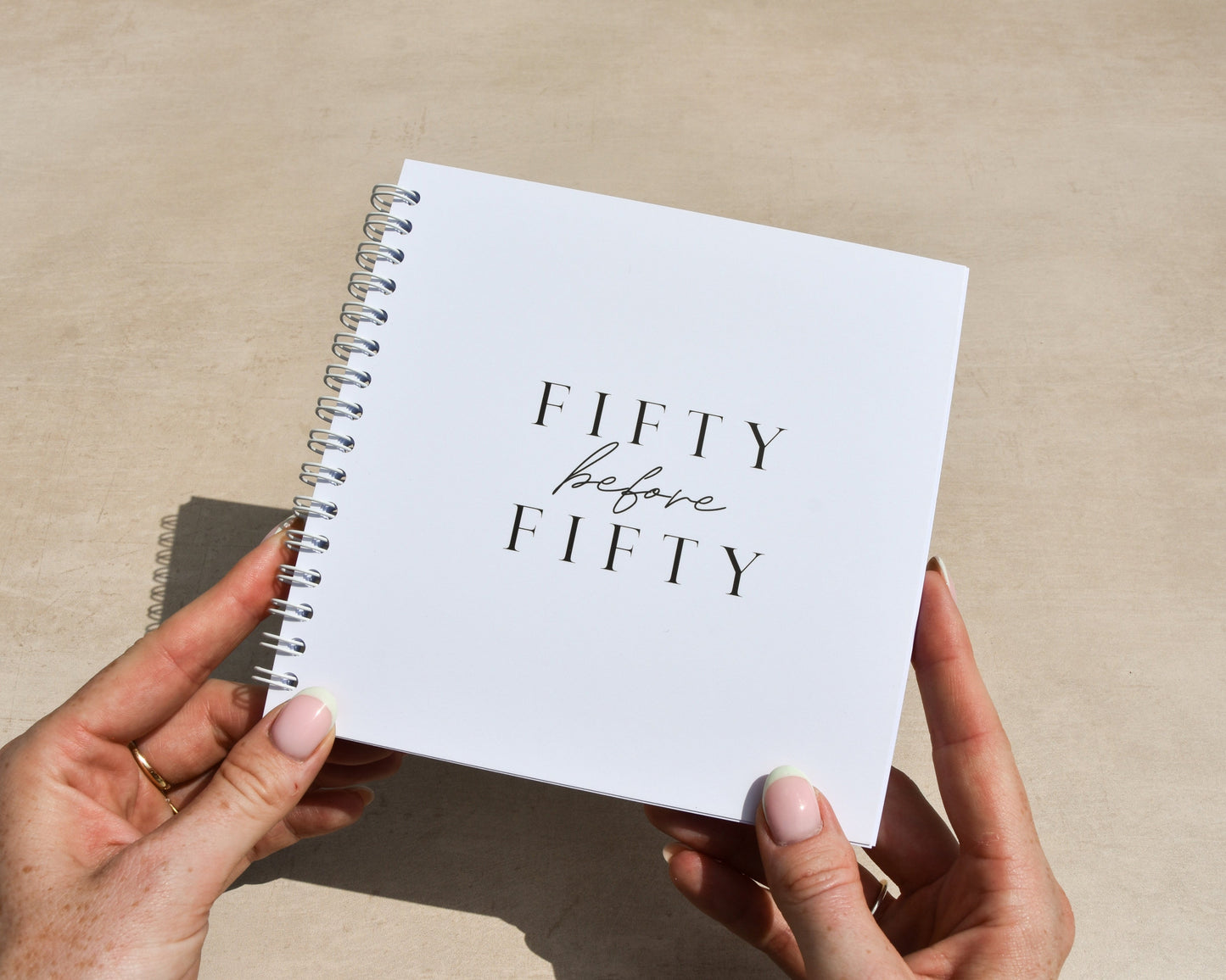 Person holding a notebook titled 'Fifty before Fifty' on a neutral background