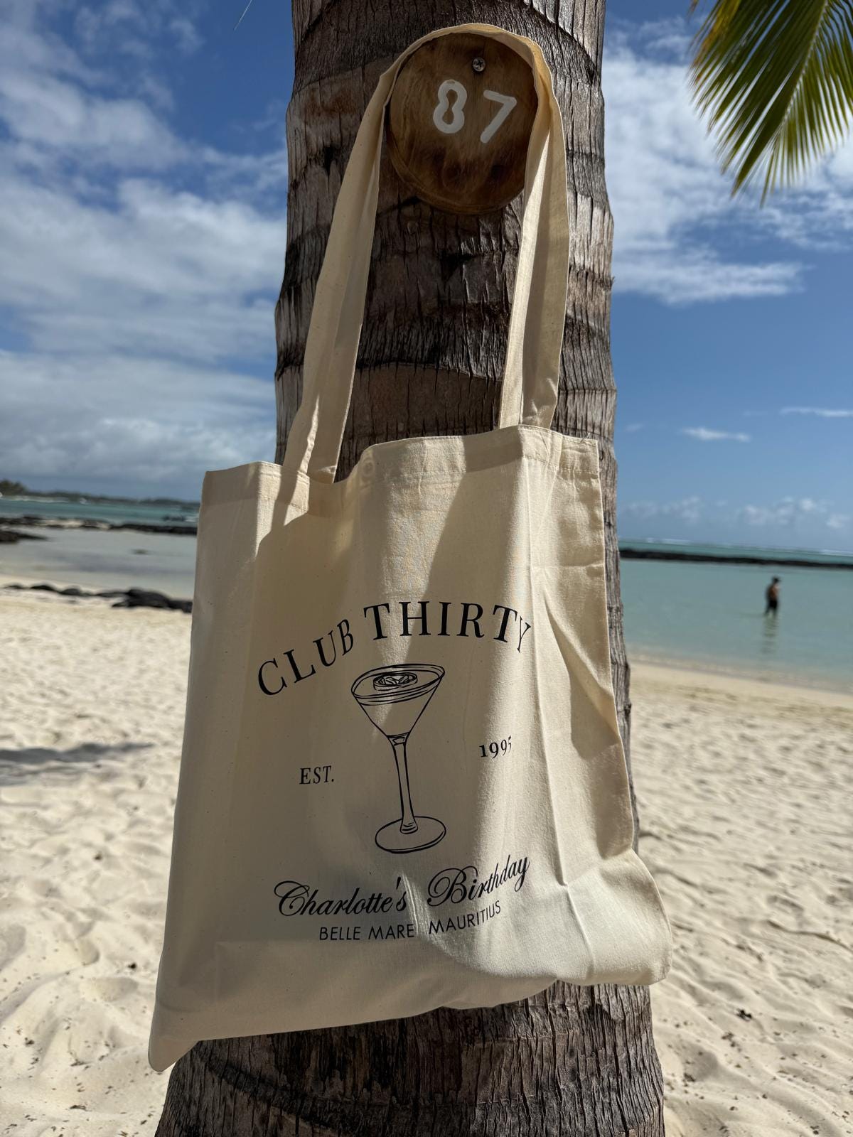Beige tote bag with 'Club Thirty' design hanging on a palm tree by the beach.