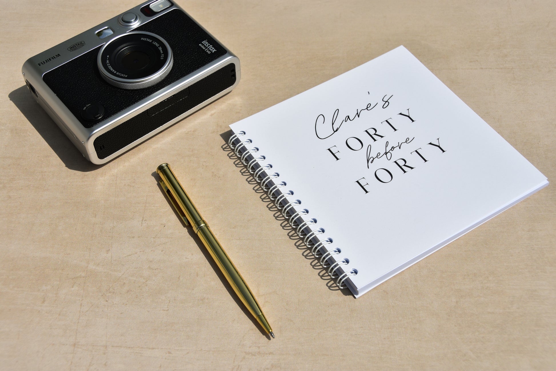 Camera, personalised 40 before 40 notebook with text, and pen on a beige surface