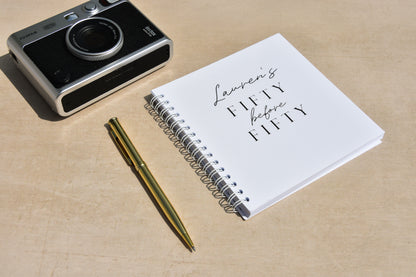 Spiral-bound notebook with 'Lauren's Fifty before Fifty' text, pen, and camera on a beige surface