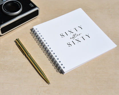 Camera, pen, and notebook titled 'Sixty after Sixty' on a wooden surface