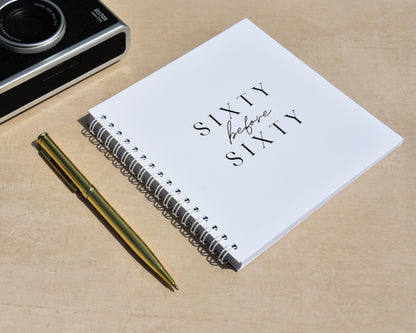 Camera, pen, and notebook with 'sixty before sixty' on a wooden surface