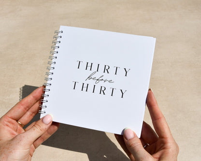 Person holding a notebook titled 'Thirty before Thirty' on a desk.